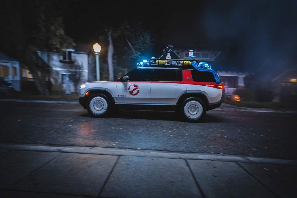 Side view of the white Rivian Ecto-R1 at night with a roof rack, red stripe, and No-Ghost logo