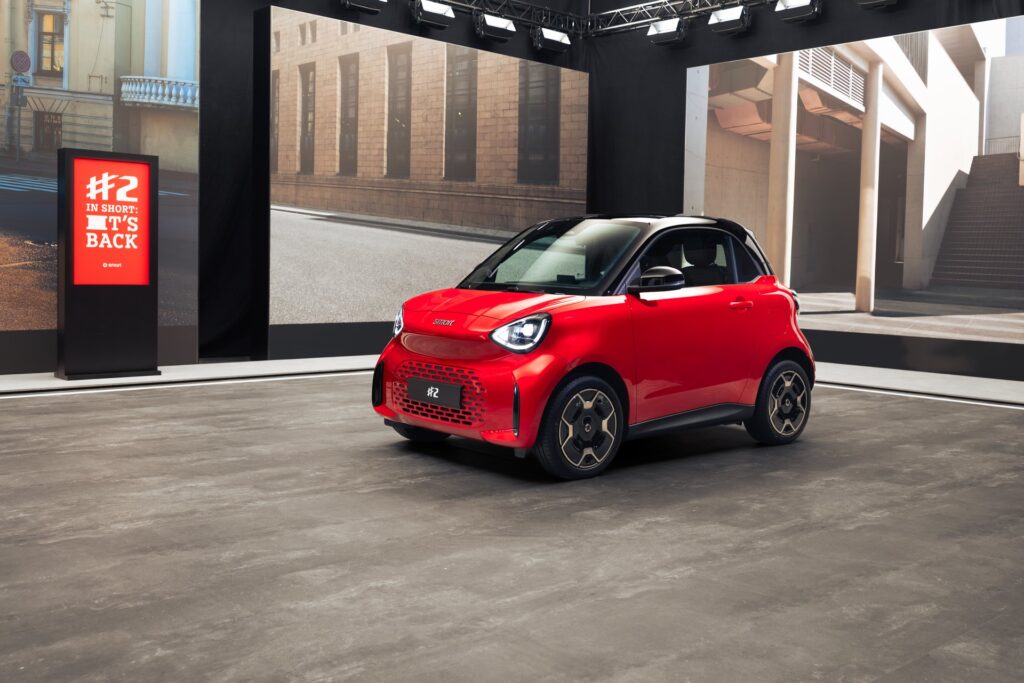 Red smart #2 two-seater on a studio stage beside an 'In short: it's back' sign
