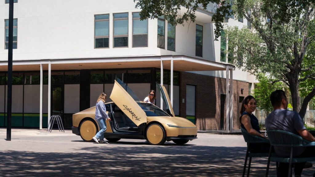 Gold Tesla Cybercab with butterfly doors open as a rider climbs in beside an outdoor seating area