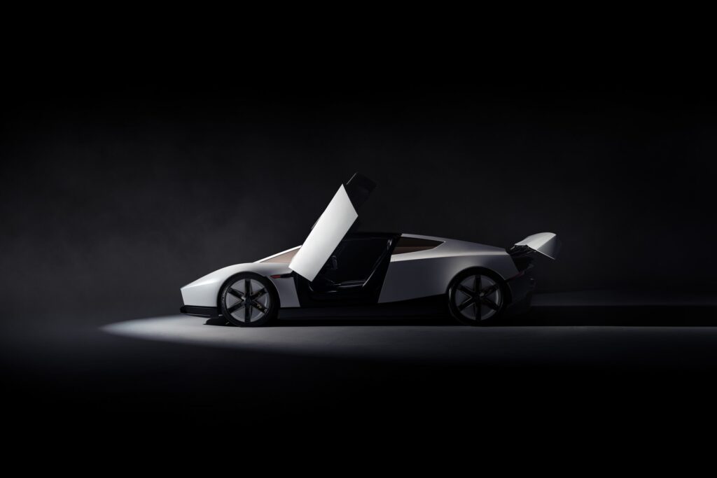 Side profile of a white Tesla Roadster with its door raised and rear wing deployed under a spotlight