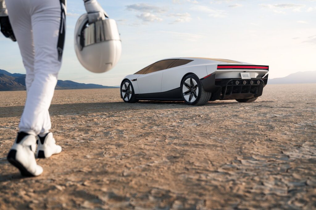 A driver in a white race suit carries a helmet toward a white Tesla Roadster on a dry lakebed