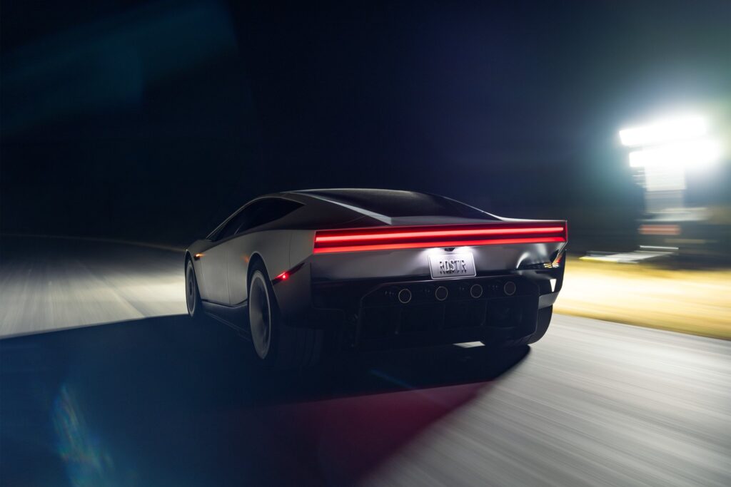 Tesla Roadster speeding along a road at night with its red light bar streaking past floodlights