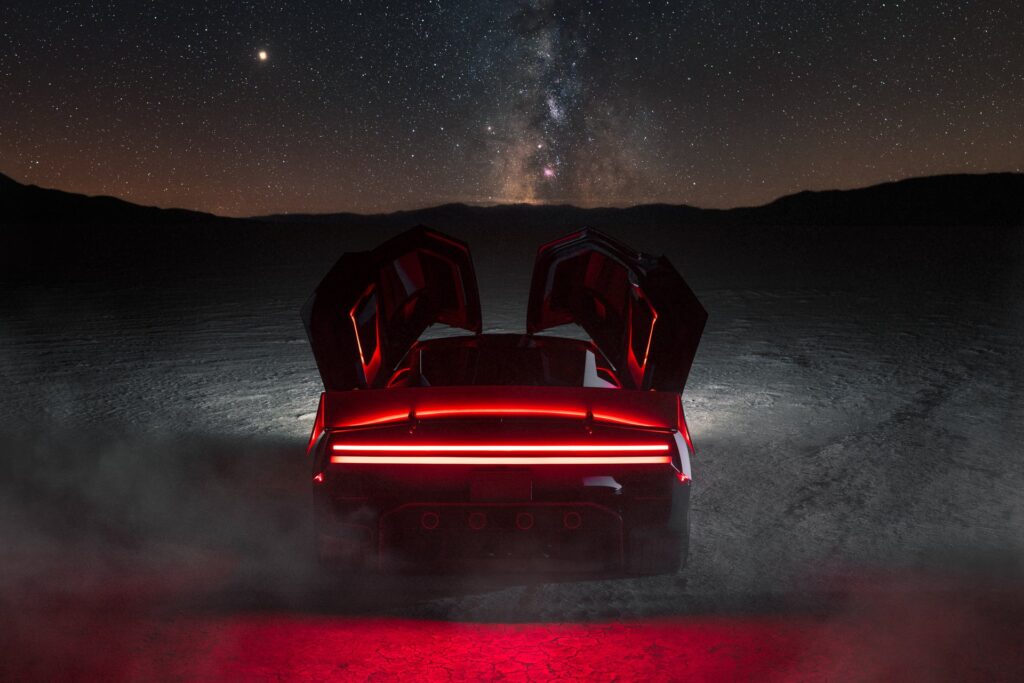 Tesla Roadster at night under the Milky Way with doors raised, its red lights glowing through dust