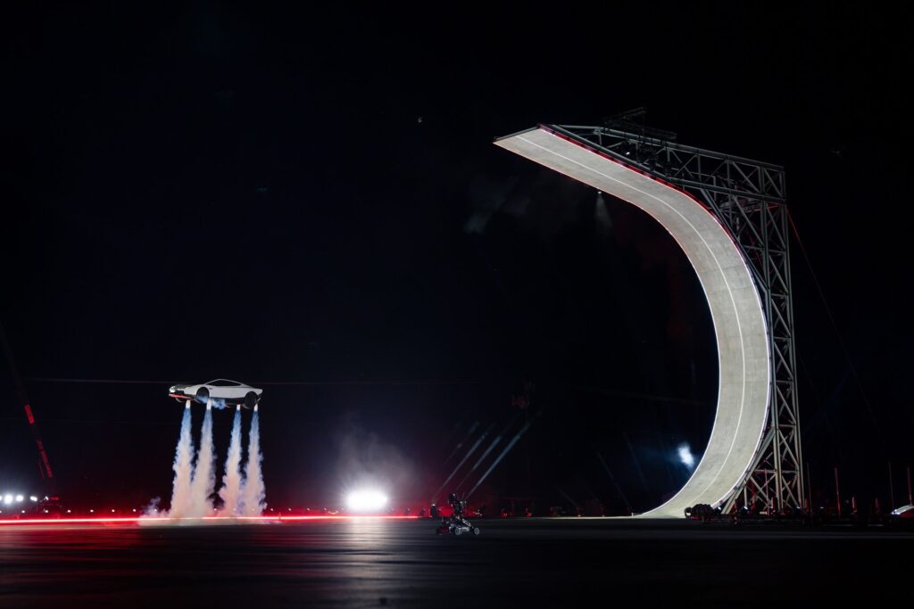 Tesla Roadster hovering on four white thruster plumes beside the C-shaped ramp at night