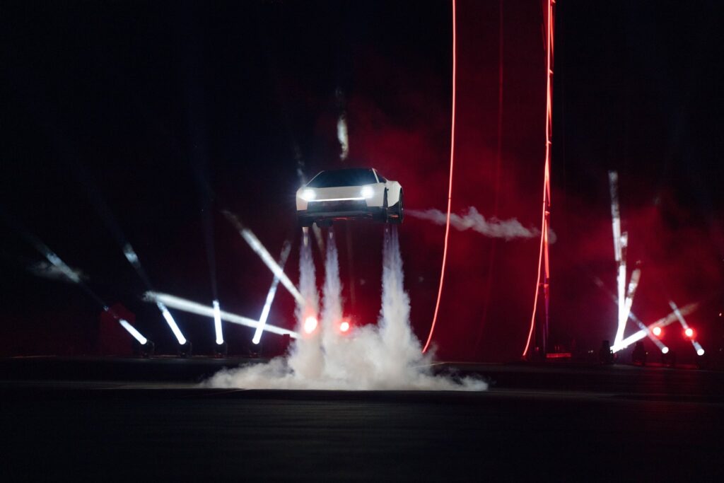 Head-on view of the Tesla Roadster hovering on thruster plumes amid red lights and stage beams