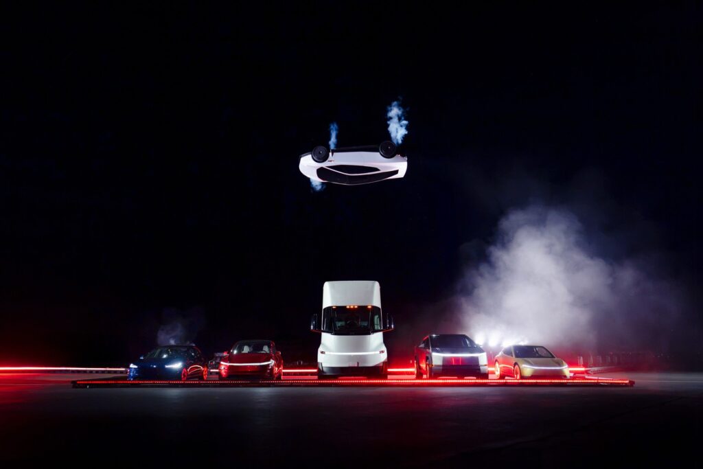 White Tesla Roadster upside down in midair above a lineup of Tesla vehicles including the Semi and Cybertruck