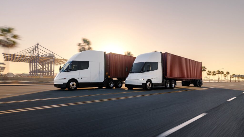Two Tesla Semi trucks hauling shipping containers near a port at sunset